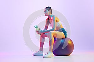 Young woman resting between trainings sitting on fit-ball with battle of water in neon light against gradient studio photo