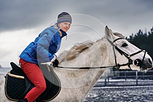 Young woman rider in a blue blazer and sporting a cap for a walk on a white horse on a cloudy winter day.