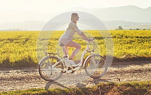 Young woman rides electric bicycle on dusty country road, view from side, sun backlight field with yellow flowers background