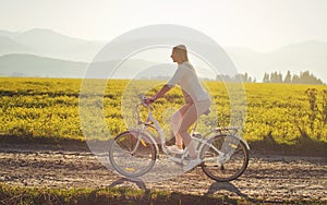 Young woman rides electric bike over dusty country road, strong afternoon sun backlight in background shines on yellow flowers