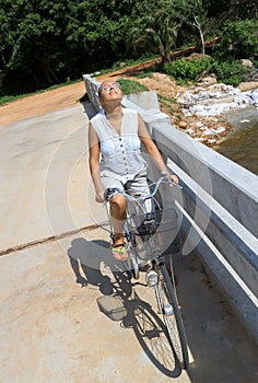 Young woman riding bicycle across river bridge next to tropical park
