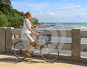 Young woman riding bicycle across river bridge next to tropical park