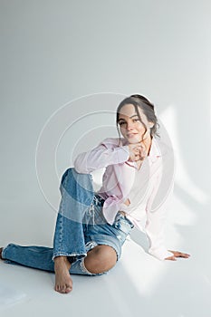 Young woman in ripped jeans and shirt poses sitting barefoot and sunlit