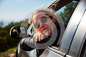 Young Woman On Road Trip Vacation Leaning Out Of Rental Car Window With Mountain Landscape Behind