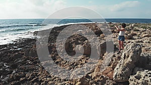 Young woman on rocks taking pictures with camera. Strong sea ocean waves hitting coastline. Aerial drone view. Sunset in cyprus