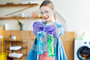 young woman in rubber gloves holding plastic bottles with cleaning fluids and smiling