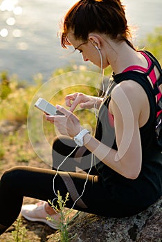 Young Woman Runner Using Smartphone and Listening to Music at Sunset on the Mountain Trail. Sports Concept