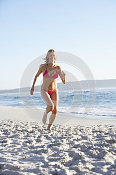 Young Woman Running Along Sandy Beach On Holiday Wearing Bikini