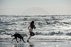 young woman running barefoot in swimsuit on the beach with her dog