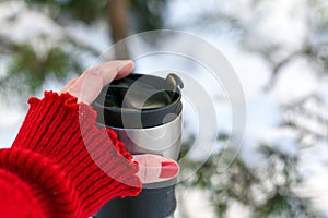 A young woman`s hand in a bright red knitted mitten holding a cup of coffee against the winter snowy forest background