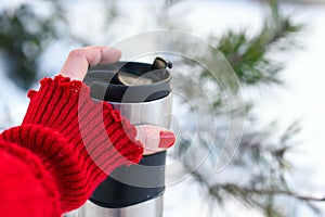 A young woman`s hand in a bright red knitted mitten holding a cup of coffee against the winter snowy forest background