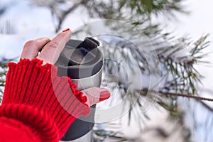 A young woman`s hand in a bright red knitted mitten holding a cup of coffee against the winter snowy forest background