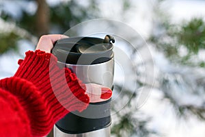 A young woman`s hand in a bright red knitted mitten holding a cup of coffee against the winter snowy forest background