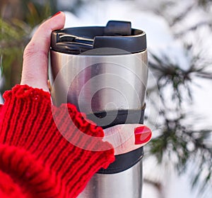 A young woman`s hand in a bright red knitted mitten holding a cup of coffee against the winter snowy forest background