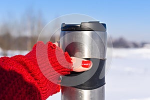 A young woman`s hand in a bright red knitted mitten holding a cup of coffee against the winter snowy forest background