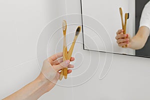 A young woman's hand holds two natural wooden bamboo toothbrushes on the background of a mirror in the bathroom.