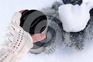 A young woman`s hand in a knitted mitten holding a cup of coffee against the winter snowy forest background