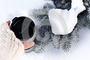 A young woman`s hand in a knitted mitten holding a cup of coffee against the winter snowy forest background