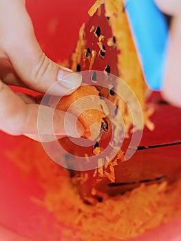 A young woman's hand rubs the carrots on a grater into a red bowl