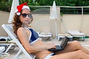 Young woman in santa claus hat working on laptop by the pool, christmas vacation in a tropical country. Woman freelance working