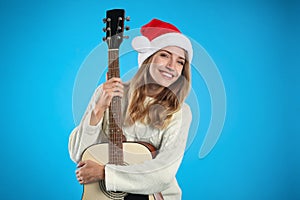 Young woman in Santa hat with acoustic guitar on light blue background. Christmas music