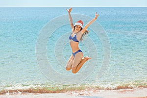 Young woman in Santa hat and bikini jumping on beach, space for text. Christmas vacation