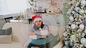 Young woman in santa hat with gift box sitting on couch near Christmas tree in living room. Happy female considers and
