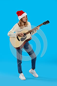 Young woman in Santa hat playing acoustic guitar on light blue background. Christmas music