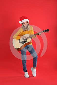 Young woman in Santa hat playing acoustic guitar on red background. Christmas music