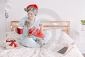 Young woman in Santa hat playing with cute puppy dog pet at home. Pet owner celebrating Christmas holiday alone. Distant remote