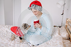 Young woman in Santa hat playing with cute puppy dog pet at home. Pet owner celebrating Christmas holiday alone. Holiday