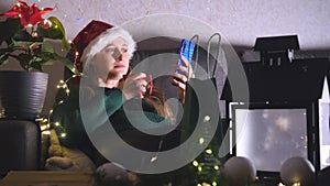 Young woman in Santa hat sitting on the sofa drinking tea and using mobile phone.