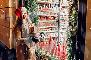 Young woman in Santa's hat holding Christmas presents gift boxes by store showcase on city street at night. Shopping