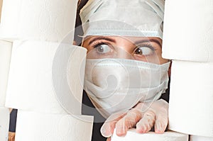 Young woman with scared eyes in two medical virus protection face masks looks through stacks of toilet paper. Covid-19 quarantine