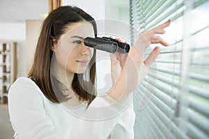 young woman searching with binoculars and looks out through blinds
