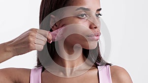 A young woman is seen using a rose quartz gua sha tool on her jawline.