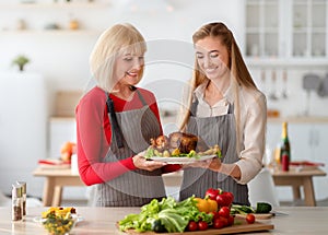 Young woman with senior mother holding dish with traditional turkey, cooking Christmas or Thanksgiving dinner in kitchen