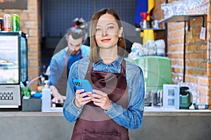 Young woman service worker in apron with smartphone in restaurant, coffee shop