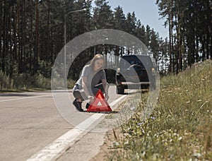 Young woman setting red triangle caution sign on road side near broken car