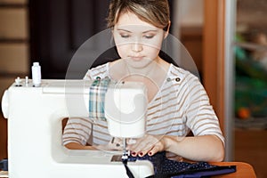 Young woman sewing with sew machine at home while sitting by her working place. Fashion designer carefully creating new