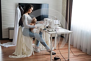 Young woman sews at home and holds a small child. Mom teaches her little son to sew on a sewing machine