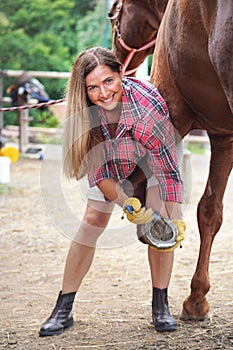 Young woman in shirt cleaning horse hoof with metal horseshoe