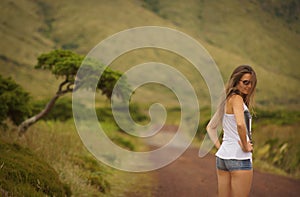 Young woman in shirt and shorts with sunglasses turning head backwards on country road. View from behind