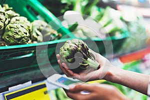 Young woman shopping healthy food in supermarket blur background. Female hands buy products artichoke using smartphone in store