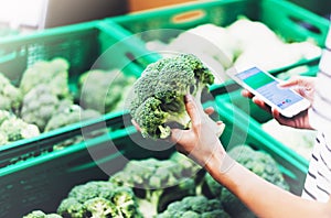 Young woman shopping healthy food in supermarket blur background. Female hands buy products broccoli using smartphone in store