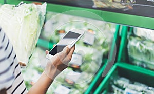 Young woman shopping healthy food in supermarket blur background. Female hands buy products cabagge using smart phone in store photo