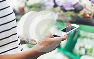 Young woman shopping healthy food in supermarket blur background. Female hands buy products cabagge using smartphone in store photo