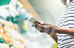 Young woman shopping healthy food in supermarket blur background. Female hands buy products tomato using smartphone in store