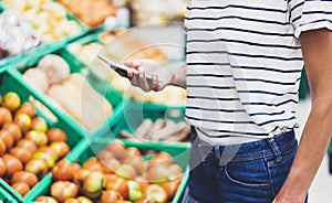 Young woman shopping healthy food in supermarket blur background. Female hands buy products tomato using smartphone in store