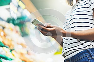 Young woman shopping healthy food in supermarket blur background. Female hands buy products tomato using smartphone in store